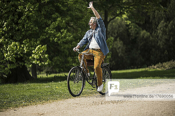 Smiling senior man riding bicycle waving hand at park on sunny day