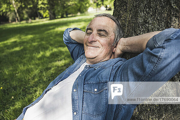 Smiling senior man with eyes closed leaning on tree trunk at park