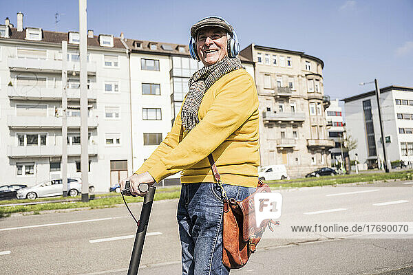 Happy senior man wearing wireless headphones riding electric push scooter