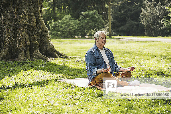 Senior man sitting cross-legged meditating on exercise mat at park