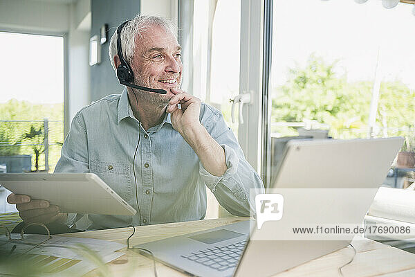 Smiling freelancer wearing headset sitting with tablet PC and laptop at home
