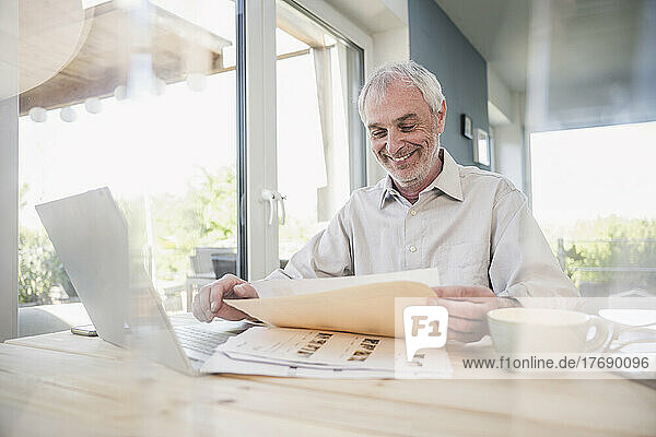 Happy senior freelancer with laptop and documents working at home