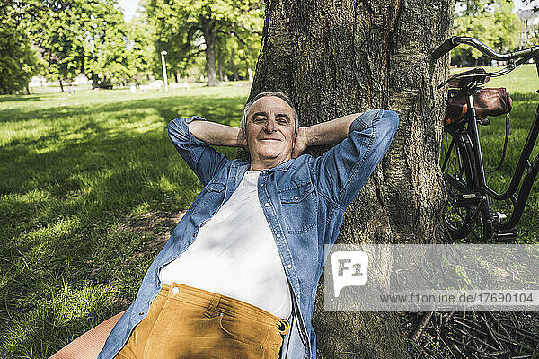 Smiling man with fully unbuttoned denim shirt leaning on tree trunk at park
