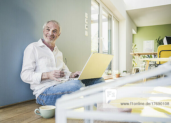 Smiling senior man with laptop and coffee cup sitting on wall at home