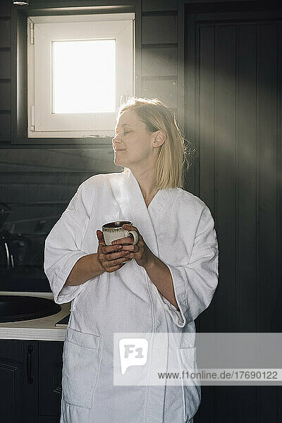 Frau im Bademantel hält Tasse in der Hand und genießt zu Hause das Sonnenlicht