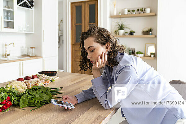 Frau benutzt Mobiltelefon neben Gemüse am Esstisch in der Küche