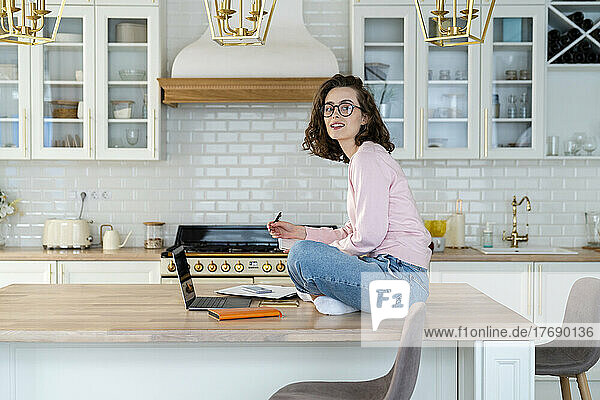 Junge Frau mit Laptop sitzt am Esstisch in der Küche
