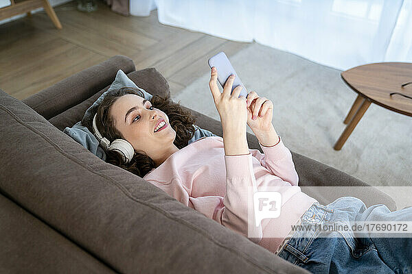 Junge Frau benutzt Mobiltelefon und liegt auf dem Sofa im Wohnzimmer
