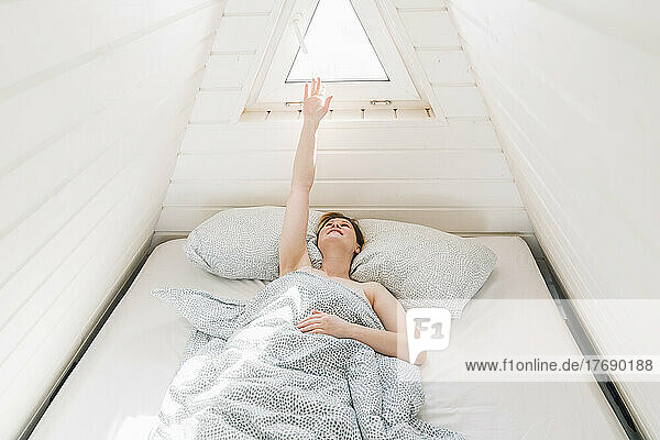 Frau mit erhobener Hand liegt auf dem Bett und genießt das Sonnenlicht im Dachboden