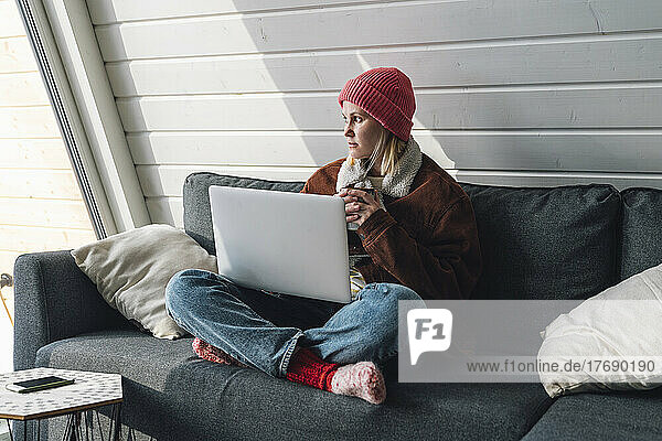 Frau in warmer Kleidung sitzt zu Hause mit gekreuzten Beinen auf Sofa und Laptop