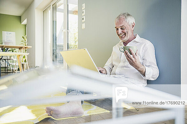 Glücklicher älterer Mann mit Kaffeetasse und Laptop  der zu Hause an der Wand lehnt