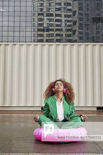 Businesswoman listening music through wireless headphones meditating sitting on pink inflatable ring