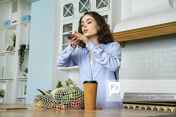 Young woman taking picture of grocery bag in kitchen