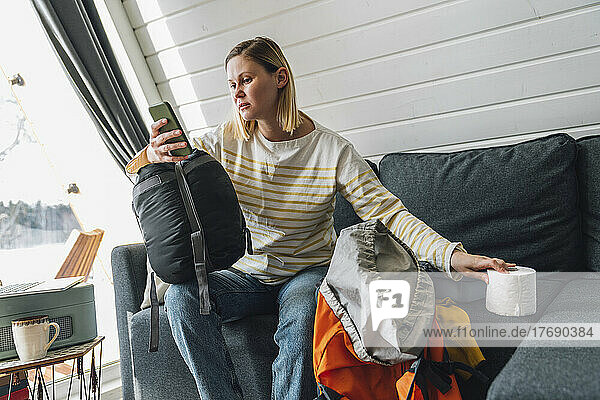 Frau benutzt Smartphone und hält Toilettenpapier in der Hand  während sie zu Hause auf dem Sofa sitzt
