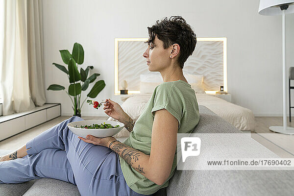 Woman eating salad with fork sitting on sofa at home