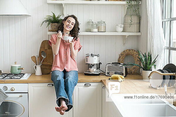 Happy woman eating breakfast sitting on kitchen counter at home