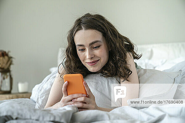 Smiling young woman using mobile phone on bed at home