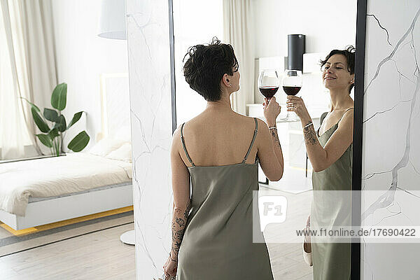 Smiling woman toasting wineglass at reflection on mirror at home