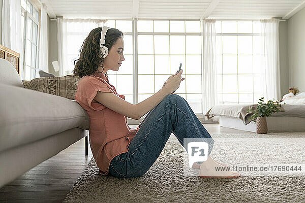 Junge Frau sitzt zu Hause auf dem Teppich und nutzt ihr Smartphone