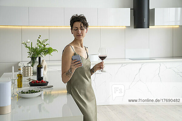Woman holding wineglass using smart phone standing in kitchen