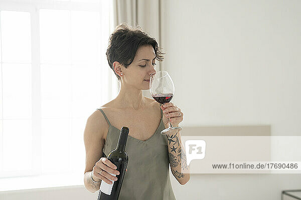Woman holding bottle smelling glass of wine standing at home