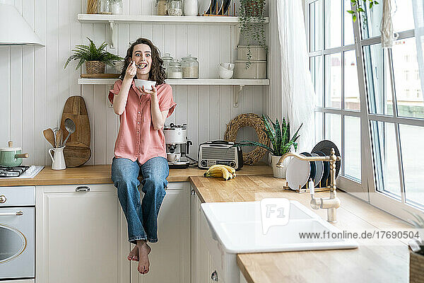 Woman eating breakfast sitting on kitchen counter at home