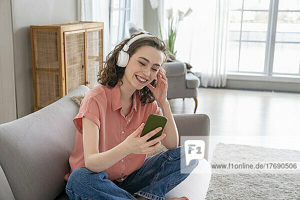 Happy woman wearing wireless headphones using smart phone on sofa at home