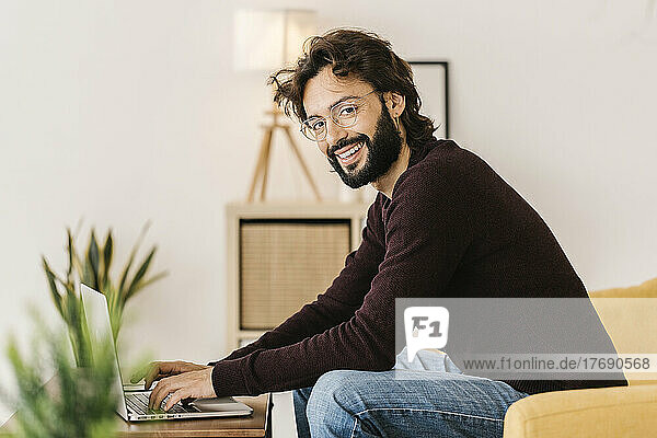 Smiling bearded hipster man with laptop sitting on sofa at home