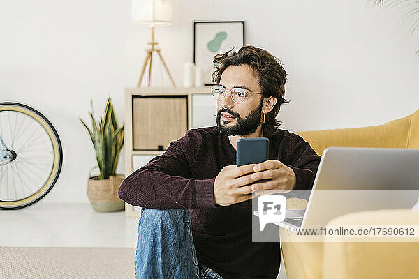 Thoughtful bearded man with mobile phone and laptop sitting by sofa at home