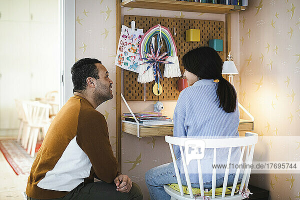 Seitenansicht des Vaters  der sich im Gespräch mit seiner Tochter  die auf einem Stuhl in der Nähe des Schreibtisches sitzt  hockt