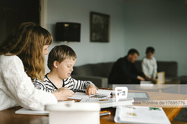 Junge mit Down-Syndrom  der von seiner Mutter ein Buch liest  während er am Tisch Hausaufgaben macht