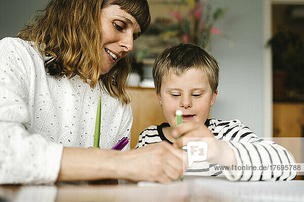 Lächelnde Mutter bringt ihrem behinderten Sohn zu Hause das Zeichnen in einem Buch bei