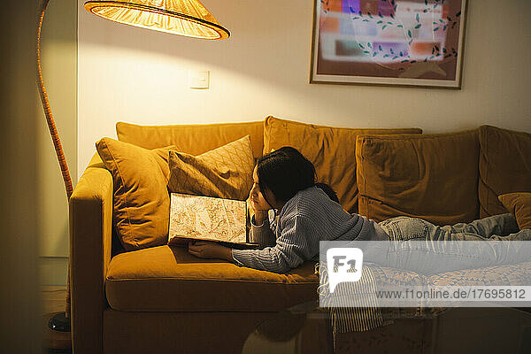 Side view of girl reading book under illuminated lamp while lying on sofa at home