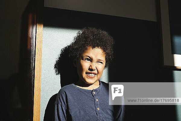 Boy winking eyes while standing in sunlight against wall at home