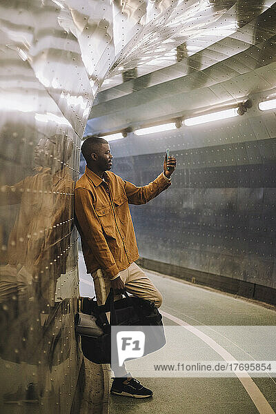 Junger Mann in voller Länge  der eine Tasche trägt  während er ein Selfie mit seinem Mobiltelefon in einem U-Bahn-Tunnel macht