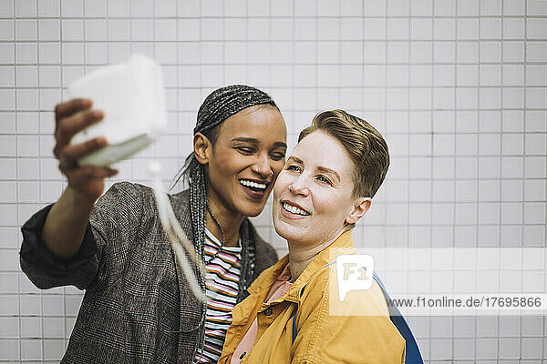 Fröhliche junge Frau  die ein Selfie mit ihrer Freundin durch die Kamera macht  während sie an der Wand steht