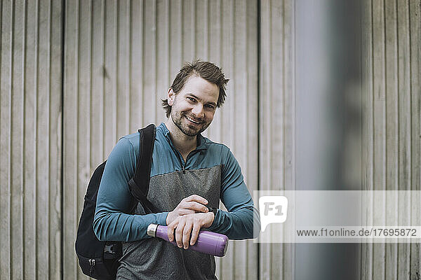 Portrait of happy young man with backpack and water bottle standing against wall