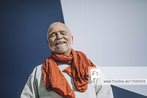 Porträt eines fröhlichen älteren Mannes mit orangefarbenem Schal  der vor einer blau-weißen Wand steht