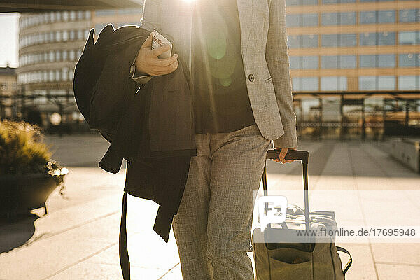 Midsection of businesswoman with smart phone pulling luggage while walking on sunny day