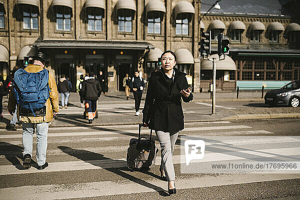 Businesswoman pulling luggage while crossing street in city on sunny day