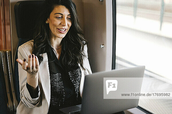 Lächelnde Geschäftsfrau führt einen Videogespräch am Laptop  während sie im Zug am Fenster sitzt