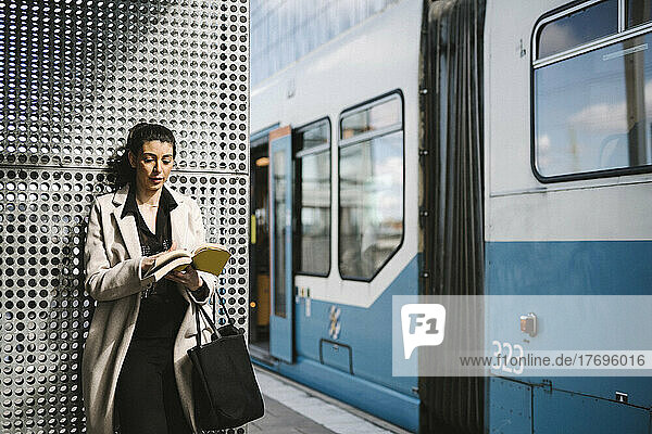 Geschäftsfrau in langem Mantel liest ein Buch beim Warten am Bahnhof
