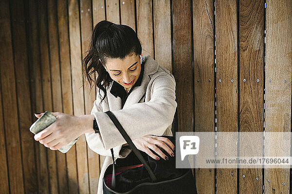 Businesswoman holding smart phone while searching in bag against wooden wall