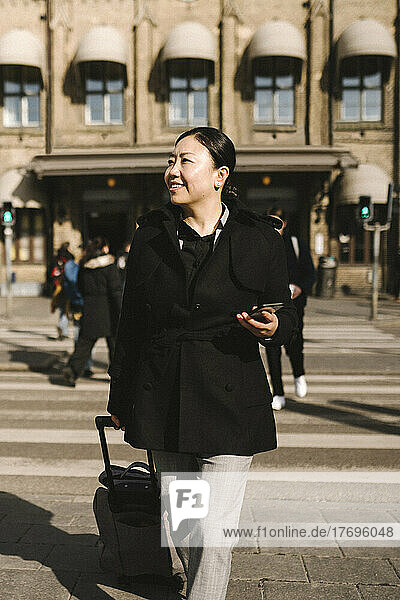 Lächelnde Geschäftsfrau mit Gepäck hält Smartphone beim Überqueren der Straße in der Stadt