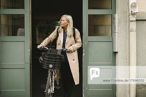 Businesswoman looking away while walking out from doorway with bicycle