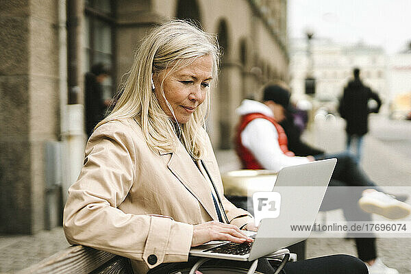 Blonde Geschäftsfrau  die einen Laptop benutzt und auf einer Bank in der Stadt sitzt