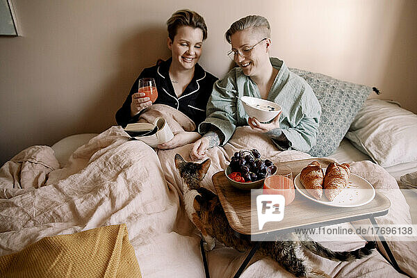 Smiling lesbian couple having breakfast in bed with cat at home