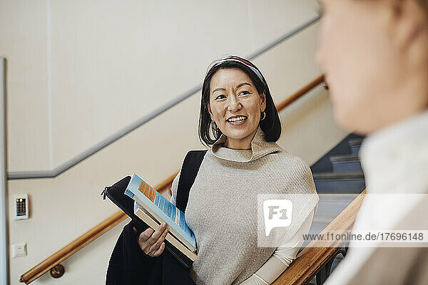 Smiling female teacher holding books while looking at colleague in university
