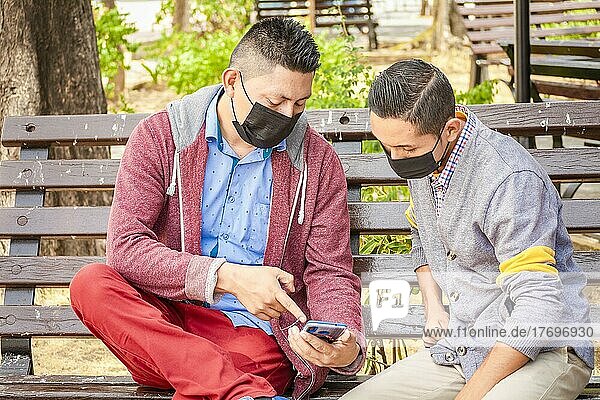Two men wearing masks checking their cell phones on a bench  a man showing his cell phone to another man