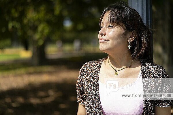 Porträt einer jungen lateinamerikanischen Frau in einem Park  die sich an einen Laternenpfahl lehnt und die Sonnenstrahlen auf ihrer Haut genießt